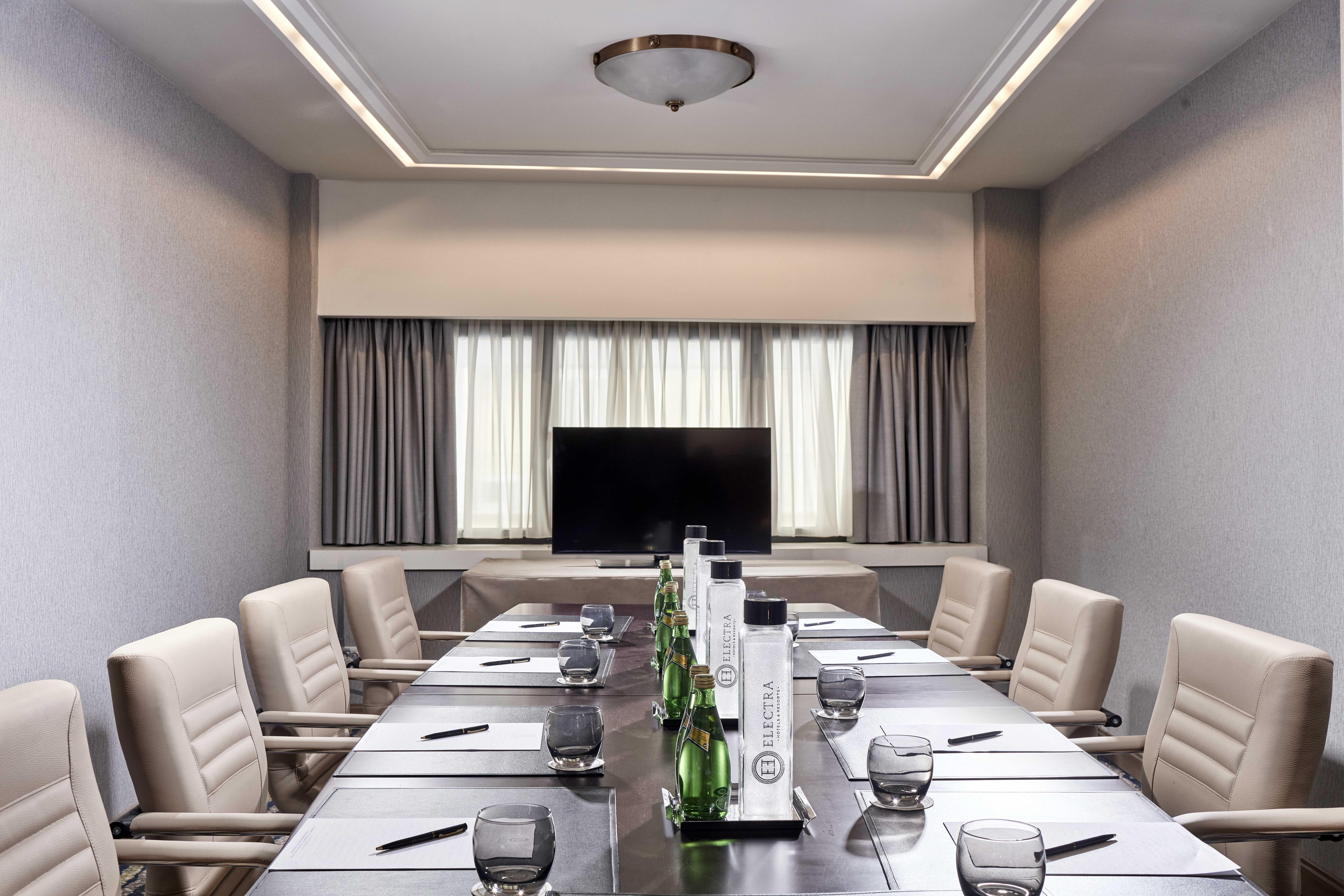 Electra Metropolis Hotel Athens Exterior photo The photo shows a modern conference room. The room features a long table surrounded by white leather chairs. On the table, there are several glass water bottles and glasses set up for attendees. A large flat-screen TV is mounted on the wall opposite 