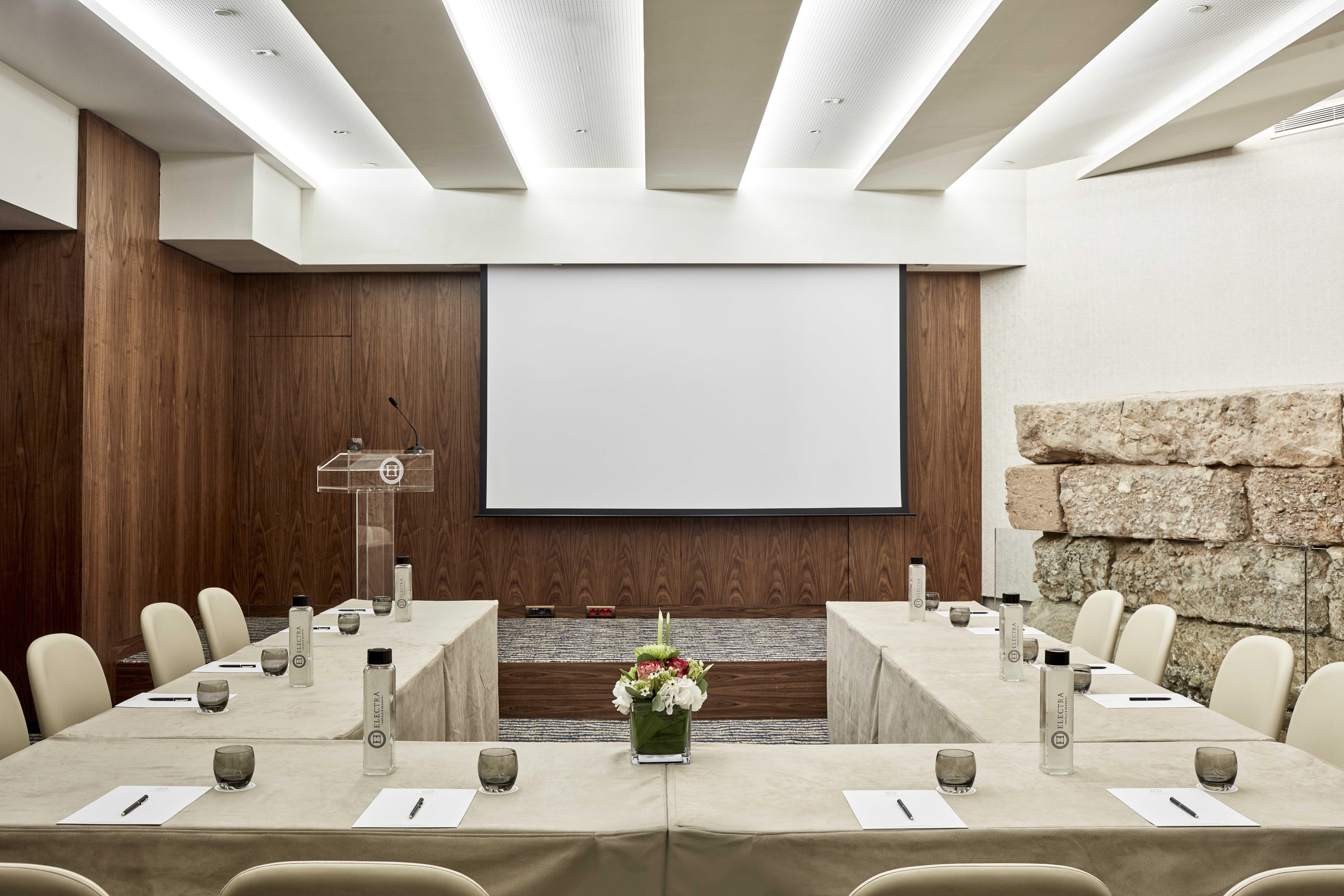 Electra Metropolis Hotel Athens Exterior photo The image shows a modern conference room setup. The room features a large screen at the front, likely for presentations. There are several tables arranged in a U-shape, covered with light-colored tablecloths, and each table has glasses and notepads. 