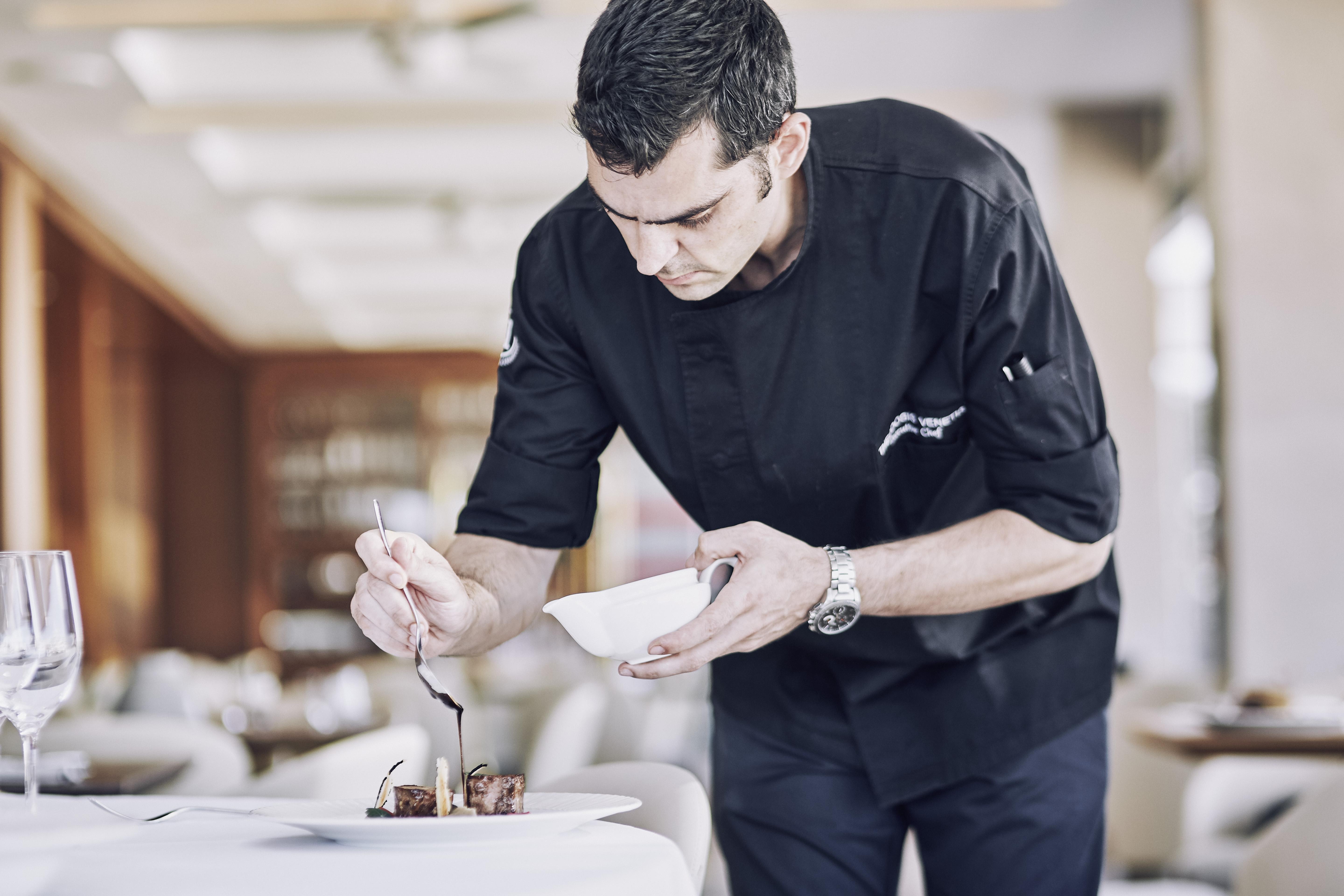 Electra Metropolis Hotel Athens Exterior photo The photo shows a chef in a black chef's jacket carefully plating a dish. He is leaning over a table, focused on adding a finishing touch to the presentation with a spoon. The dining area has a modern and elegant ambiance, with a soft focus on the ba