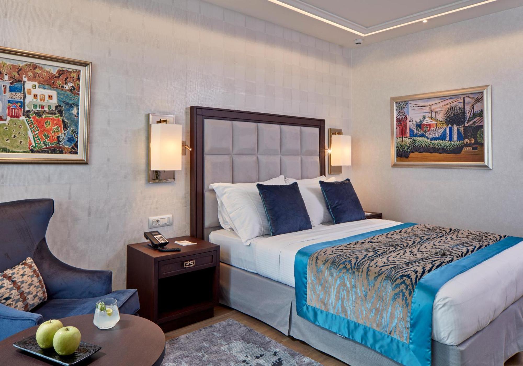 Electra Metropolis Hotel Athens Exterior photo The photo shows a modern hotel room featuring a double bed with decorative pillows. The bed has a stylish headboard, and the bedding includes a patterned throw. Beside the bed, there are bedside lamps and a wooden nightstand with a phone. 

On the le
