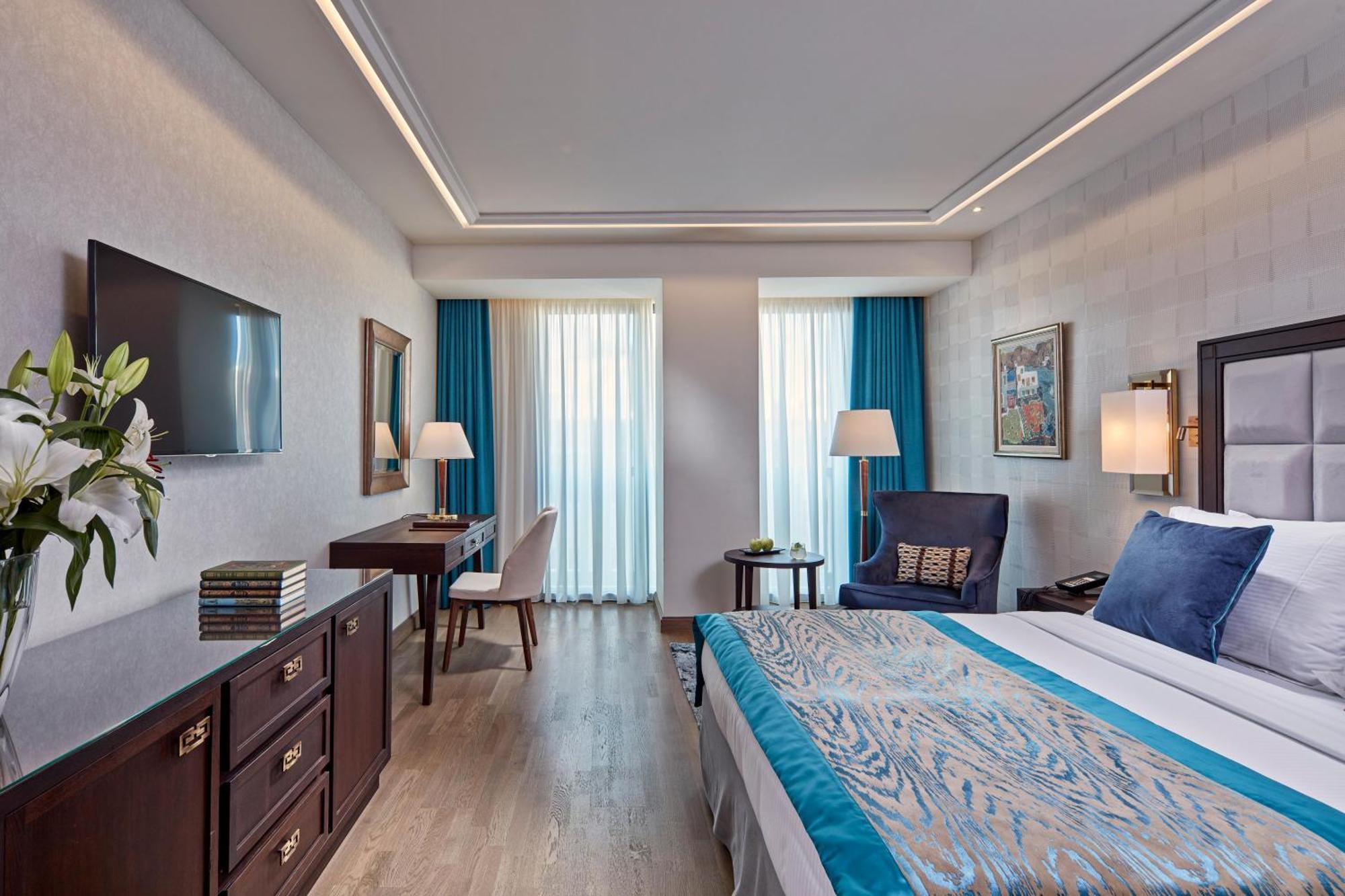 Electra Metropolis Hotel Athens Exterior photo The photo shows a well-decorated hotel room featuring a large bed with a blue and patterned bedspread. There are two windows with light blue curtains that allow natural light to fill the space. A flat-screen TV is mounted on the wall opposite the bed