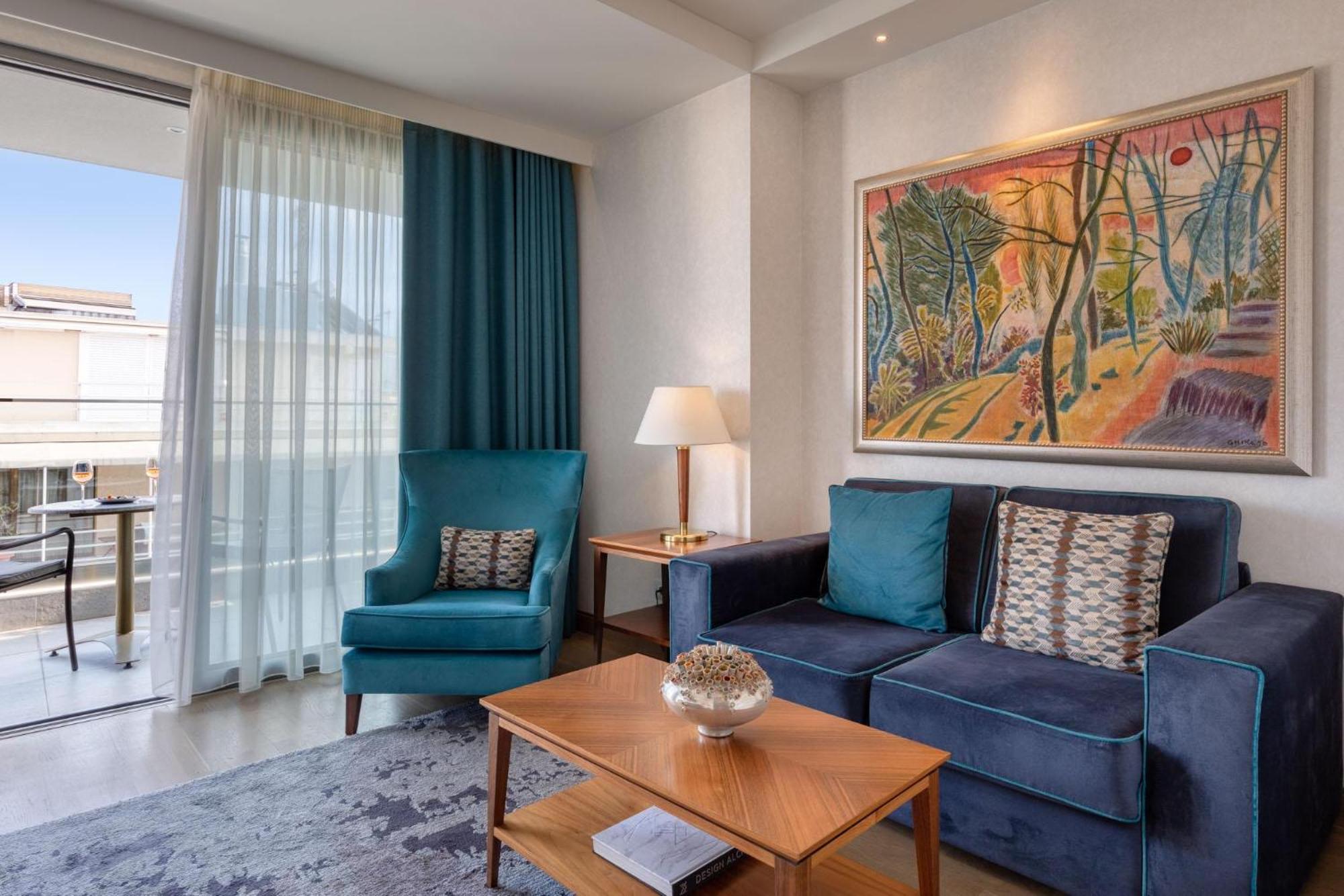 Electra Metropolis Hotel Athens Exterior photo The photo shows a modern living room area. There is a blue sofa with decorative pillows and a wooden coffee table in front of it. A light blue armchair is positioned next to the sofa. A painting with vibrant colors depicting a natural scene is hangin