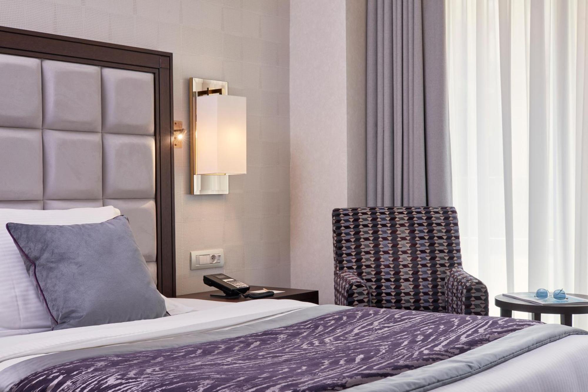 Electra Metropolis Hotel Athens Exterior photo The photo shows a modern hotel room featuring a neatly made bed with a dark purple throw blanket and large pillows. Behind the bed, there is a stylish headboard with a tufted design. A soft, ambient light fixture is mounted on the wall beside the bed