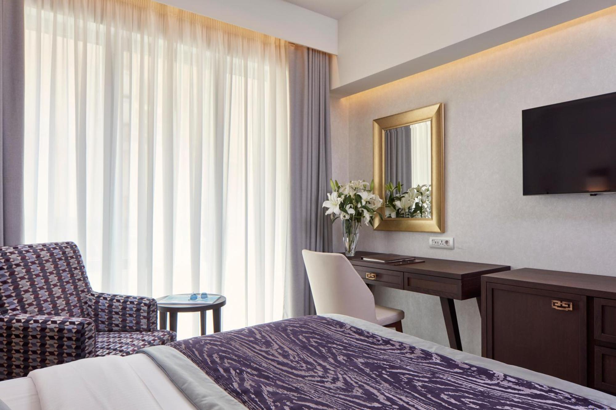 Electra Metropolis Hotel Athens Exterior photo The photo shows a well-furnished hotel room. On the left, there is a patterned chair with a small round table next to it. The room features large windows covered with sheer curtains that allow natural light to fill the space. In the center, there is 