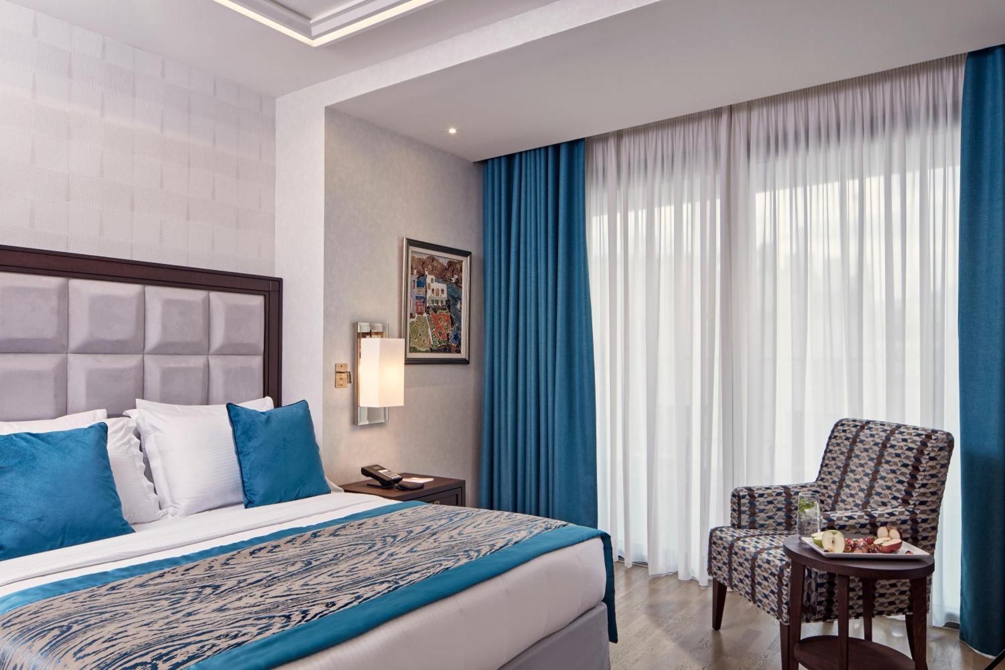 Electra Metropolis Hotel Athens Exterior photo The photo shows a modern hotel room featuring a neatly made bed with blue and white accents in the bedding. There is a textured headboard behind the bed and a piece of wall art hanging nearby. Natural light filters in through large, sheer curtains, a