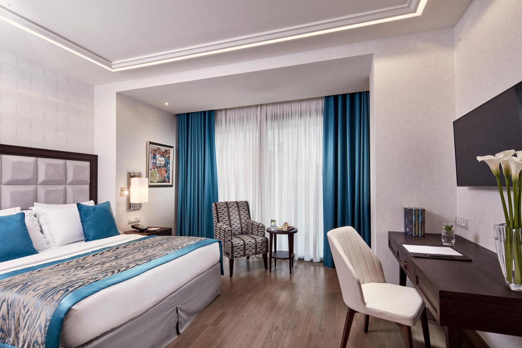 Electra Metropolis Hotel Athens Exterior photo The photo shows a well-decorated hotel room. It features a large bed with a blue and neutral colored bedding set. There are two blue curtains that frame a window, allowing natural light to enter the space. Next to the bed, there's a small chair and t