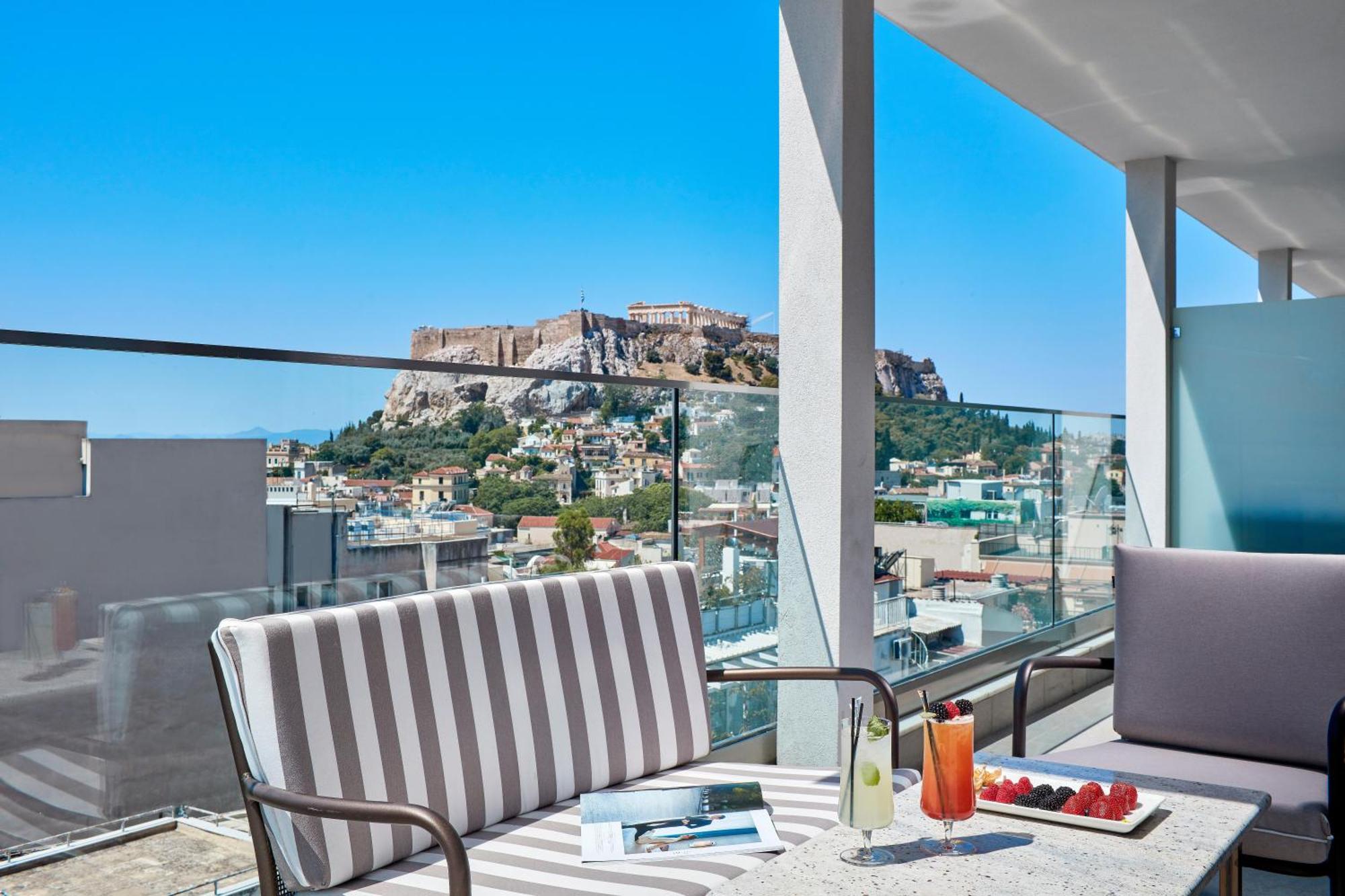 Electra Metropolis Hotel Athens Exterior photo The photo features a modern terrace or balcony with stylish seating arrangements. There are two cushioned chairs, one with striped fabric. A table in front holds a beverage, possibly a cocktail, and some fresh strawberries. The backdrop showcases a b