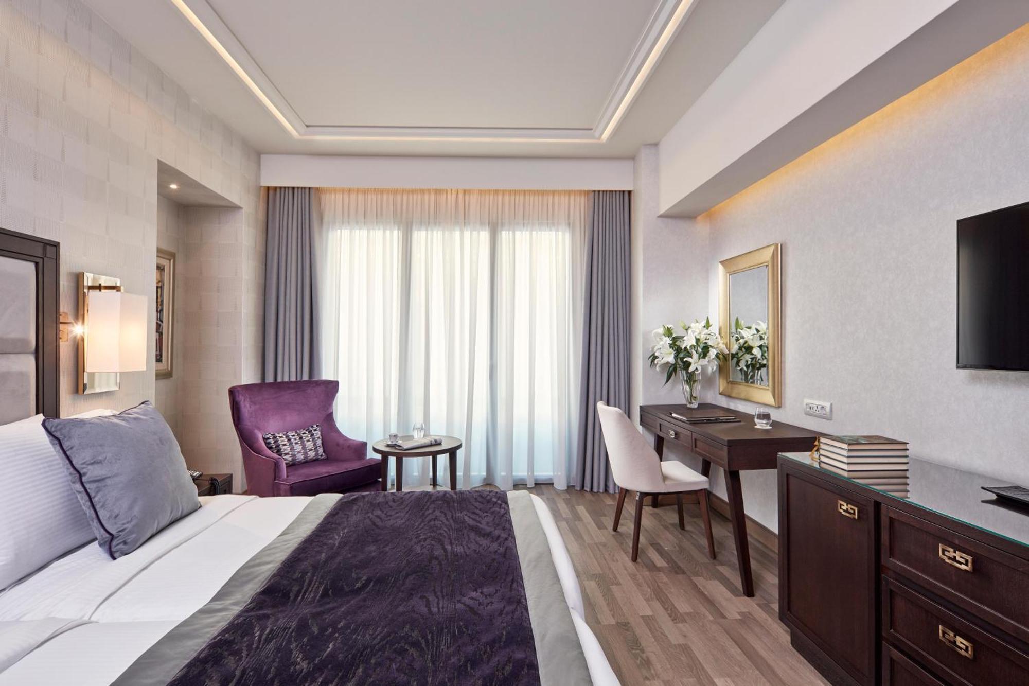 Electra Metropolis Hotel Athens Exterior photo The photo shows a well-decorated hotel room with a modern and elegant design. The room features a large bed with a dark purple throw blanket and plush pillows. There is a cozy purple armchair beside a small round table. A stylish white chair is posit