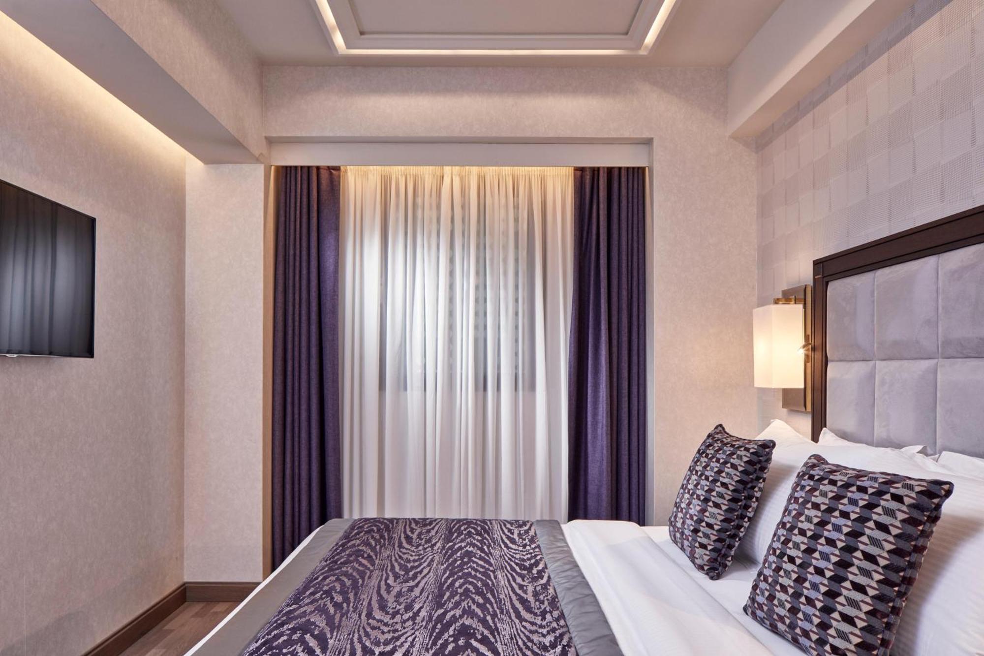 Electra Metropolis Hotel Athens Exterior photo The photo shows a modern bedroom featuring a bed with patterned pillows and a decorative comforter. The room has soft lighting, with a wall-mounted TV visible. There are large curtains that allow natural light to filter in, and the walls have a subtl