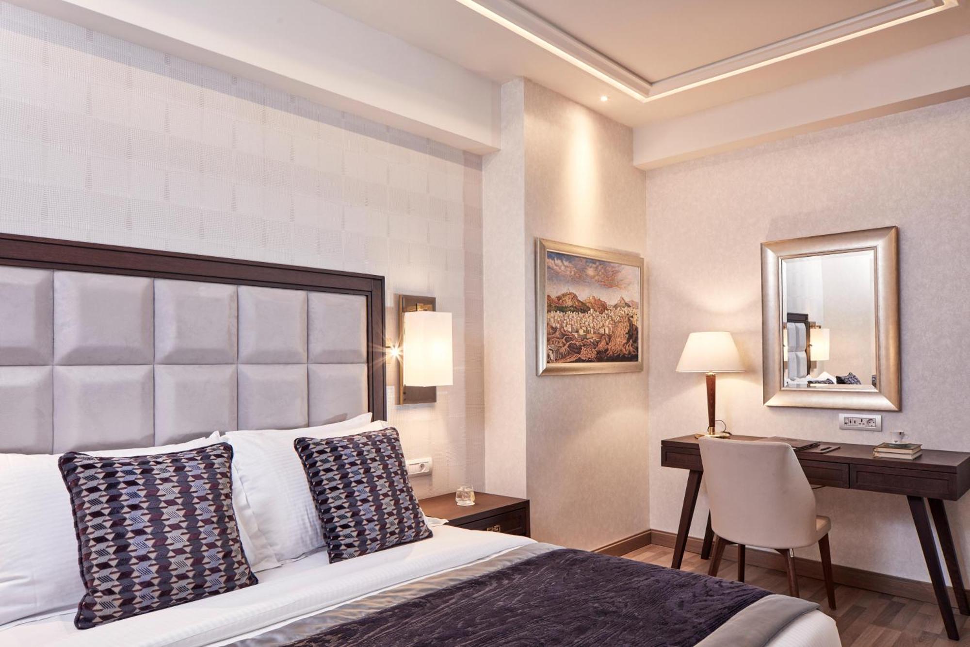 Electra Metropolis Hotel Athens Exterior photo The photo shows a modern hotel room featuring a comfortable bed with decorative pillows and a stylish headboard. The walls are light-colored with a textured design, and there are two bedside lamps providing warm lighting. To one side of the room, the