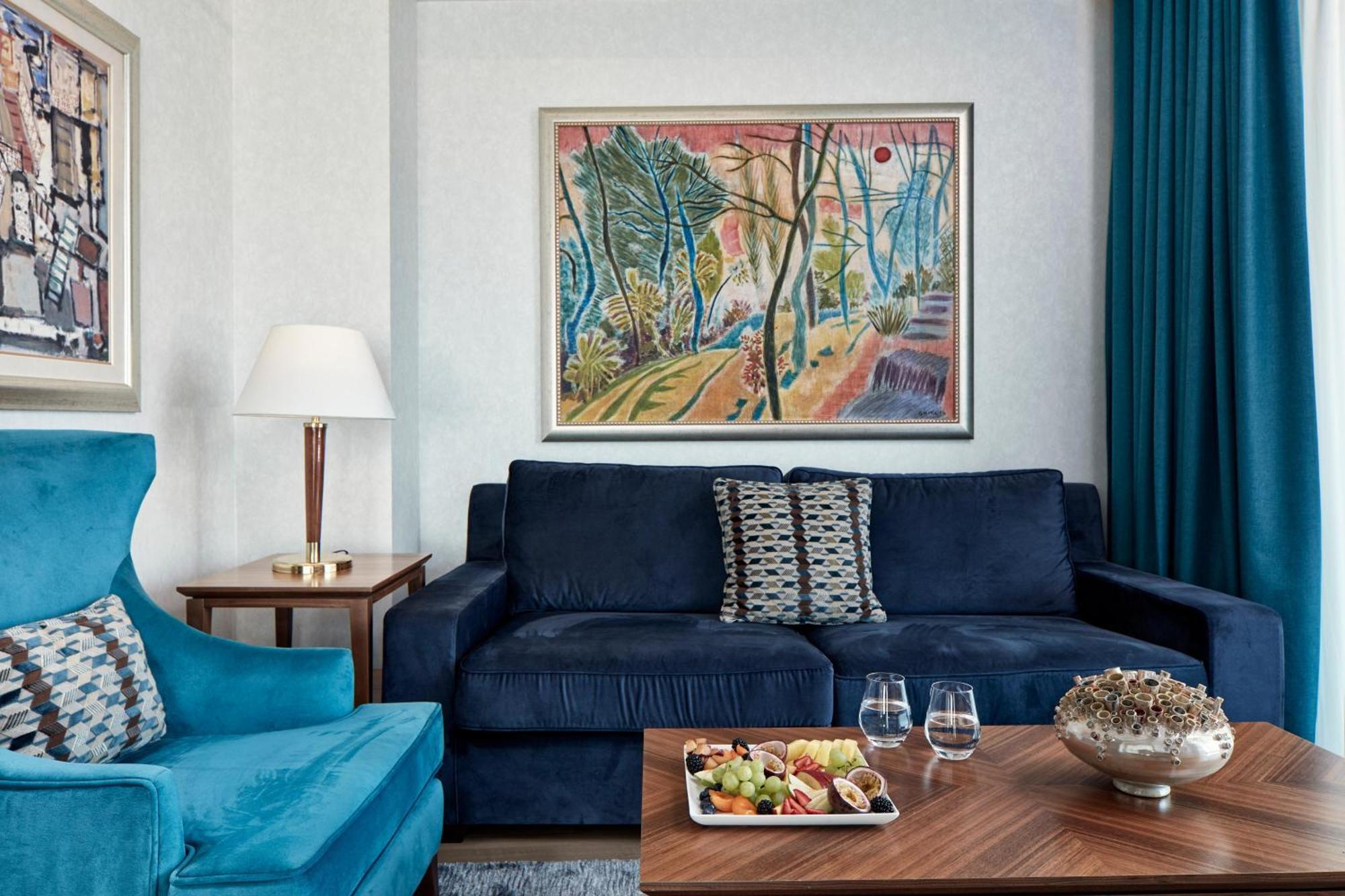 Electra Metropolis Hotel Athens Exterior photo The photo depicts a modern living room featuring a blue sofa with a decorative cushion in the center. On the left, there's a bright blue armchair and a wooden side table beside it. A lamp with a white shade is positioned on the table. The wall behind