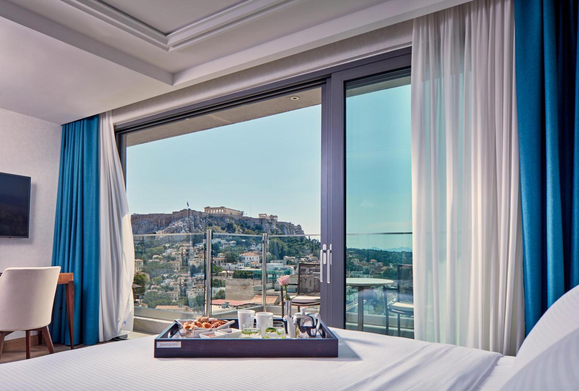 Electra Metropolis Hotel Athens Exterior photo The photo shows a modern hotel room with a large window offering a view of a historical site, likely a fortress or ancient ruins, situated on a hill. The interior features a neatly made bed with a tray placed on it, displaying various items that coul