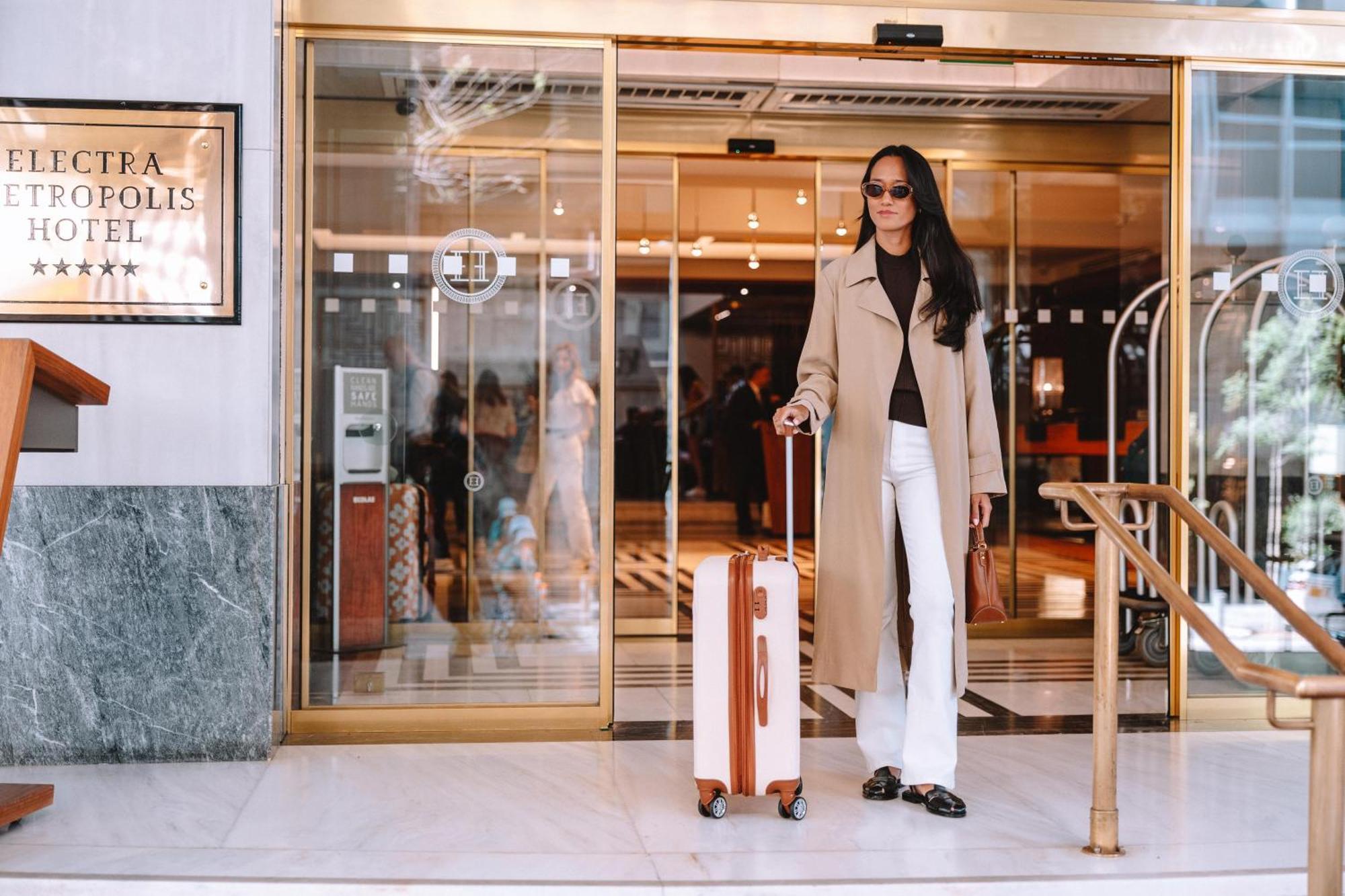 Electra Metropolis Hotel Athens Exterior photo The photo shows a fashionable individual standing outside a hotel entrance. The person is wearing a long beige coat over a dark top, paired with white pants. They are holding a stylish suitcase with a wooden design. The hotel has large glass doors, a