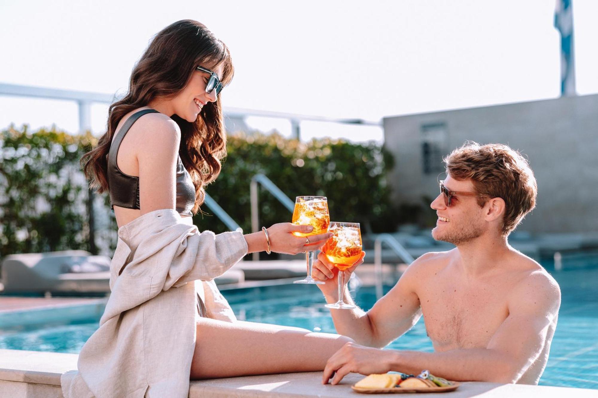 Electra Metropolis Hotel Athens Exterior photo The photo shows a pair enjoying a vibrant moment by a pool. A woman, sitting on the edge, is wearing stylish sunglasses and a bikini, and she is handing a drink to a man sitting beside her. He is shirtless, also wearing sunglasses, and is smiling whi