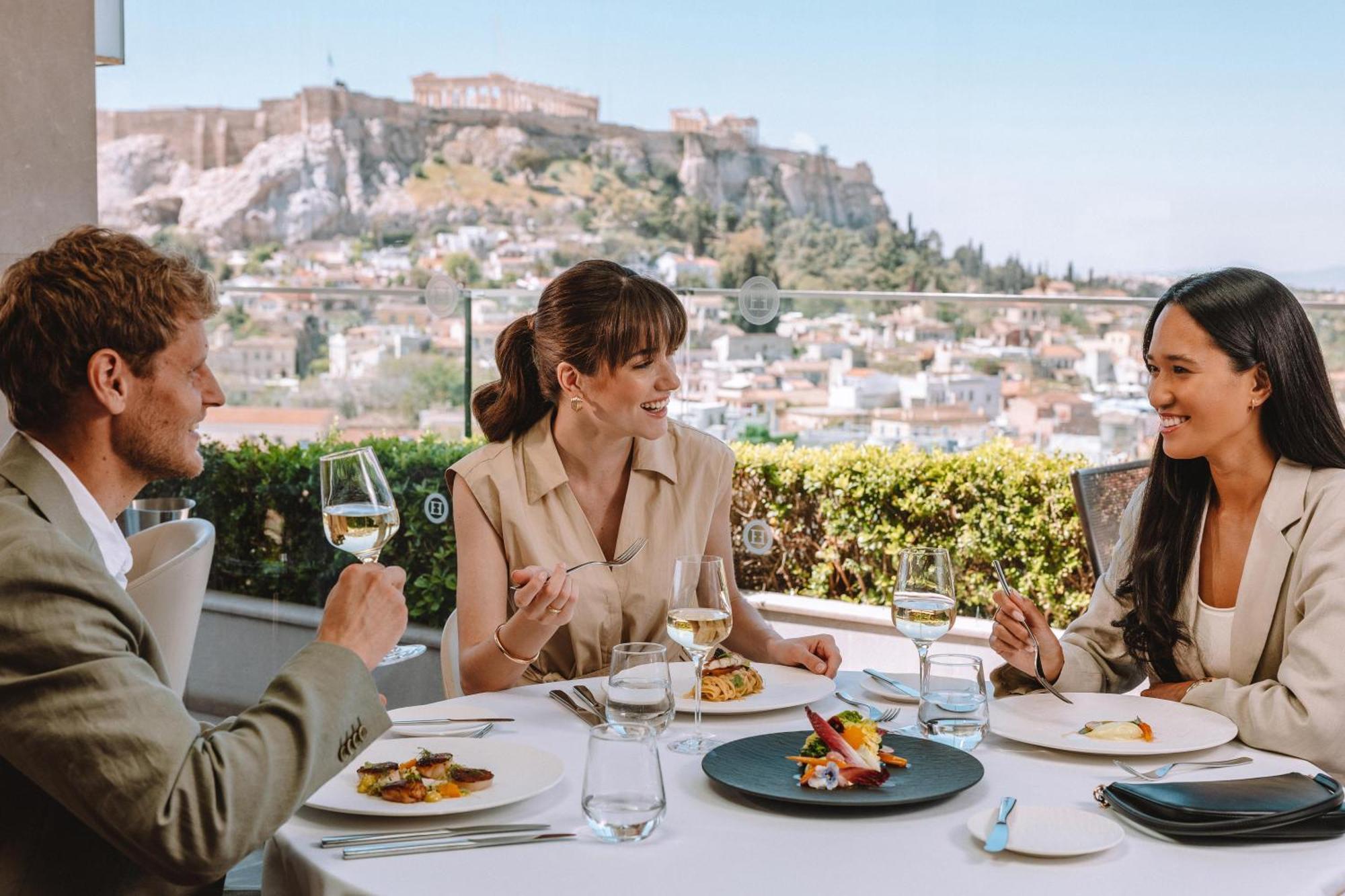 Electra Metropolis Hotel Athens Exterior photo The photo depicts three people enjoying a meal at an outdoor restaurant with a scenic view in the background. There are two women and one man seated at a table that includes several plates of food and glasses of wine. The atmosphere appears relaxed a